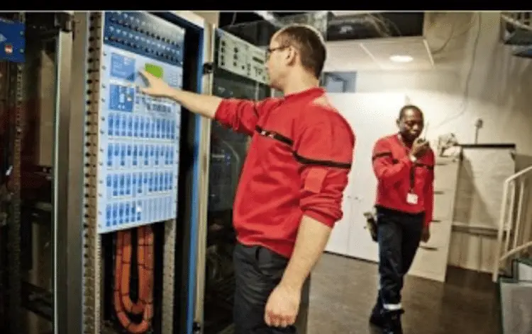 Techniciens en uniforme rouge travaillant dans une salle de serveurs, ajustant des panneaux de contrôle